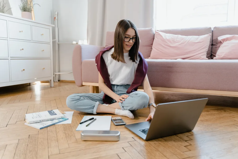 A teenage student sitting on the floor of a bright living room, studying with a laptop and notes
