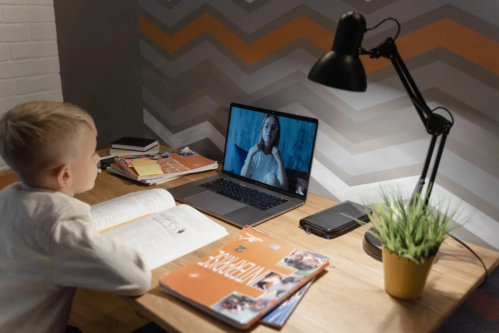 A young boy attending a live online class at a home desk, with a lamp and study books nearby