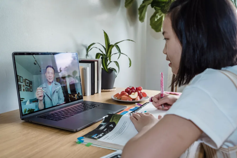 A student on a video call with a teacher, taking notes at a home desk